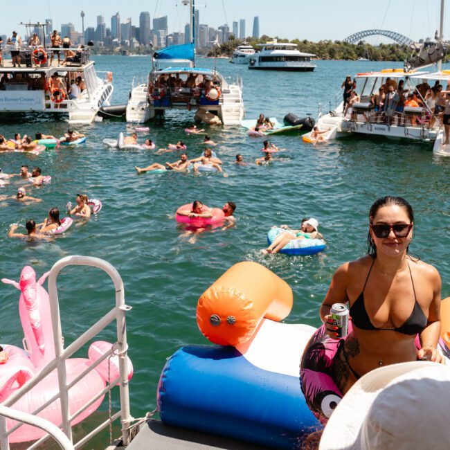 People enjoying a sunny day on the water with floaties and inflatables near several docked boats. The skyline and Sydney Harbour Bridge are visible in the background. One woman in a black bikini and sunglasses stands in the foreground holding a drink. The event is branded as "Sail Gustavo: Summer Fest.
