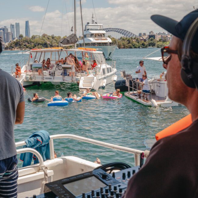 A DJ is playing music on a boat while people relax and swim in the water nearby, with other boats in the background. The lively setting features a scenic city skyline, a historic bridge, and a sunny, partly cloudy sky.