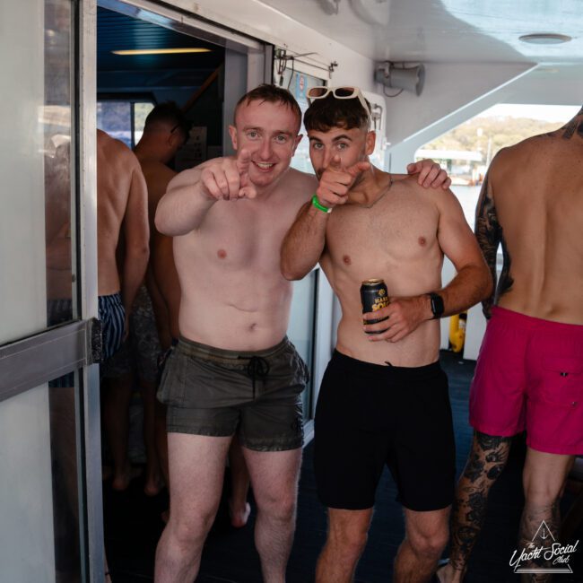 Two shirtless men in swim trunks are standing on a boat, pointing at the camera and smiling. The man on the left wears grey shorts, and the man on the right wears black shorts and sunglasses while holding a can of beer. Other people enjoying themselves are in the background.