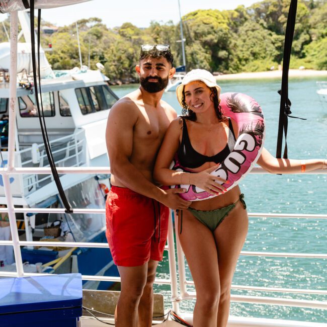 A man in red swim trunks and snorkel mask stands next to a woman in a bikini holding a pink floatation ring on a boat. The picturesque background features another boat and lush green trees by the scenic waterfront. The woman smiles while the man has a neutral expression, ready for adventure.