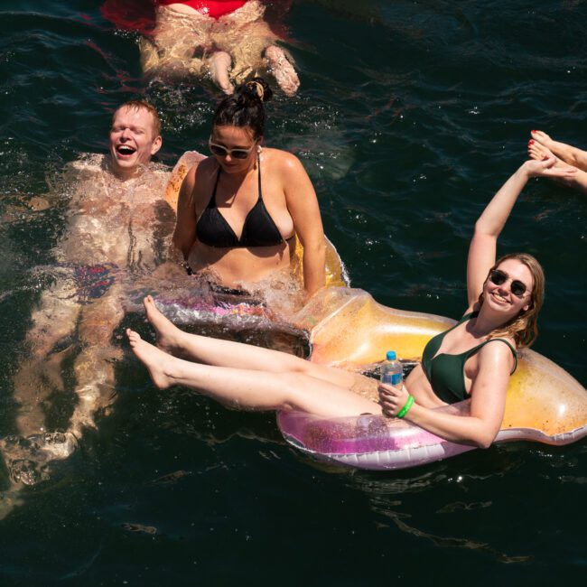 Three adults are enjoying their time in the water. One man and one woman are partially submerged, smiling and laughing. Another woman in sunglasses is relaxing on a seashell-shaped float, holding a drink and raising her arm, while another person’s legs dangle playfully in the background.