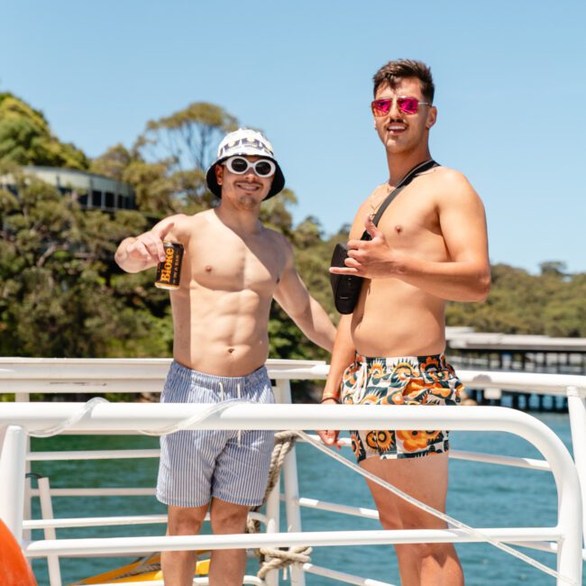 Two young men in swim trunks and sunglasses stand on a boat. One holds a can, the other forms a peace sign with his hand. The background features water, trees, and a distant structure. One man wears a bucket hat with floral patterns, while the other sports a crossbody bag.