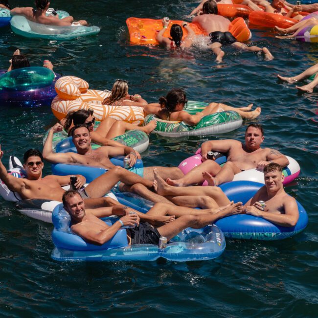 A group of people relax on various colorful inflatable floats in a beautiful, sunny body of water. Five men in swim trunks sit or recline on floats in the foreground, smiling and making hand gestures. Other individuals on floats can be seen enjoying the pleasant scene in the background.