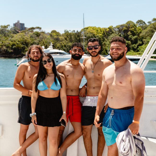 Five people stand together on a boat with water and trees in the background. Four men are shirtless, while one woman wears a black skirt and blue bikini top. They appear relaxed, posing for the photo. A logo in the corner reads "Sail Gustavo." The scene exudes summertime joy aboard a peaceful vessel.