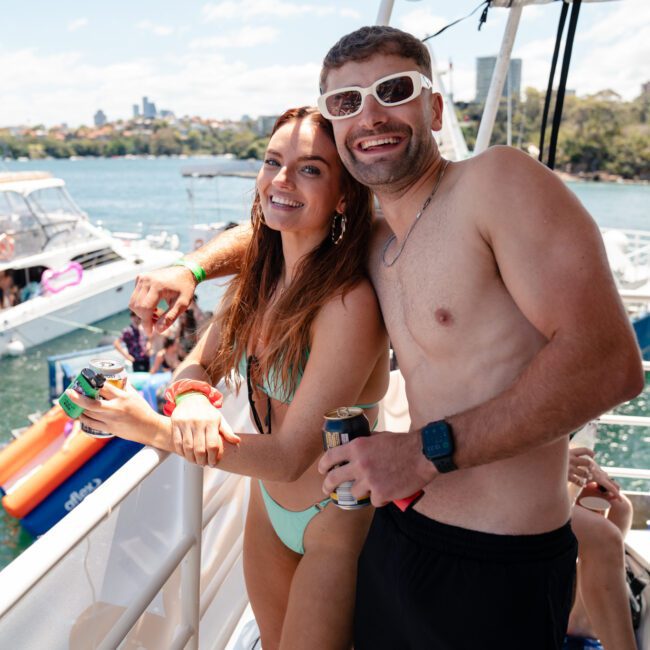Two people standing on a boat's deck, smiling at the camera during a Sydney boat party hire. The person on the left is wearing a mint-green swimsuit, while the person on the right is shirtless, wearing dark shorts and white sunglasses.