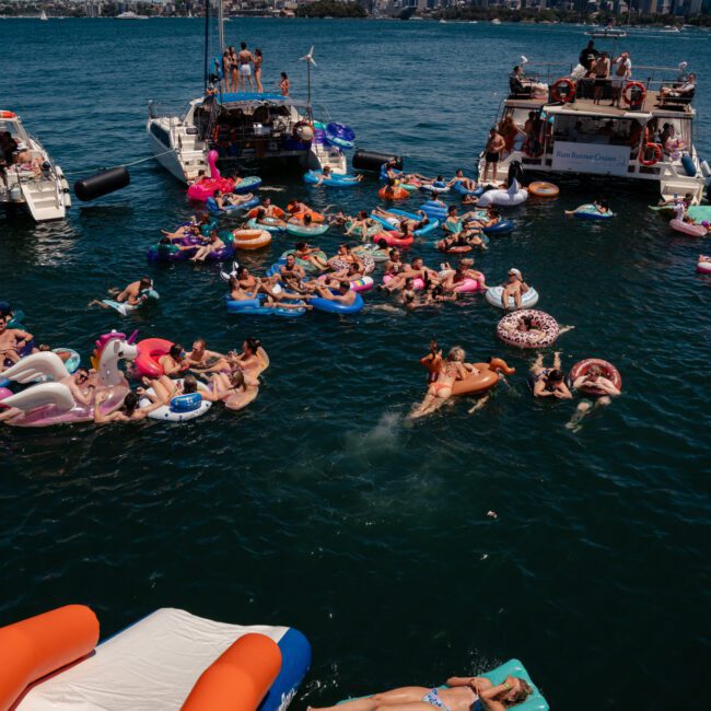 People enjoying a sunny day on boats and various flotation devices in a body of water, with a city skyline visible in the background. Consider adding to the experience with a luxury yacht hire Sydney for an unforgettable adventure.