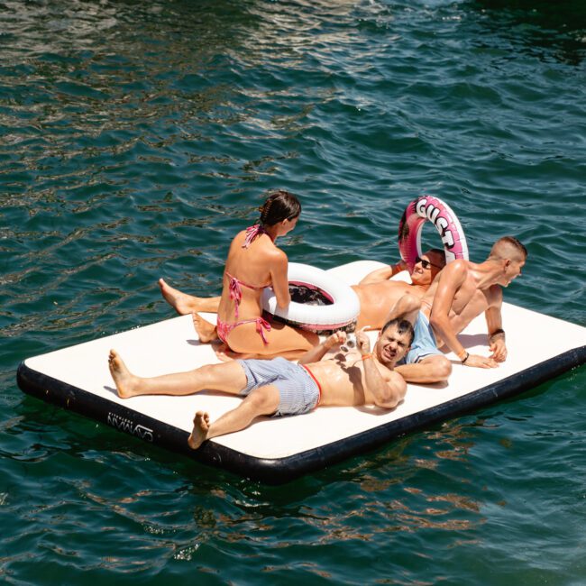 Four people are relaxing on a large inflatable platform on the water. One person in a pink bikini holds an inner tube ring with "Guac" on it. A boat filled with more people is visible in the background. The image features the logos for "Sail Gustavo" and "Utah Social.