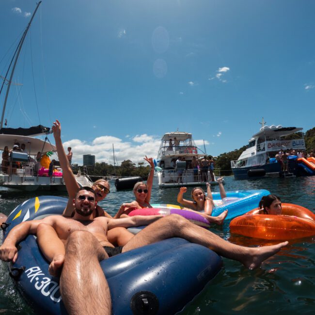 A group of people floats on inflatable rafts in a sunny, picturesque bay. Several boats are anchored in the background. The group appears to be socializing and enjoying the water, with some people waving and lounging leisurely on the colorful rafts.
