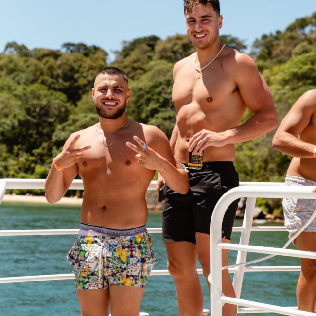Two shirtless men enjoy a sunny day on a boat, one posing with peace signs and the other holding a drink. They're surrounded by water and lush greenery in the background, both appearing happy and relaxed in their swim trunks. The "Sail Gustavo" logo is visible in the corner, enhancing their adventure.