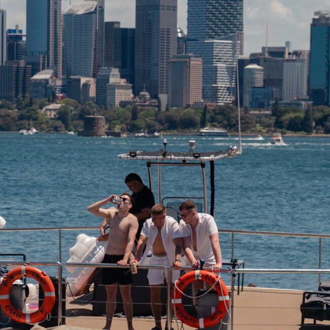 A group of people stand on the deck of a boat, enjoying the cityscape featuring tall buildings and skyscrapers in the background across the water. Two orange life rings are attached to the railing, suggesting it might be a Private yacht charter Sydney Harbour experience.