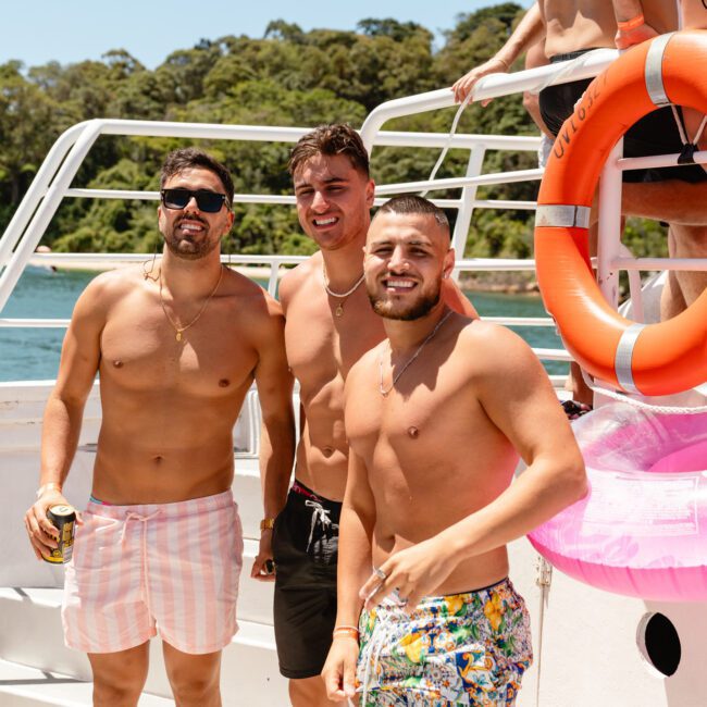 Three men in shorts stand on a boat, smiling at the camera. The man on the left wears sunglasses and holds a drink. A life preserver and another partygoer are visible in the background, with trees and a bright sky also in view. The text "SAIL GUSTAVO" is on the bottom left.