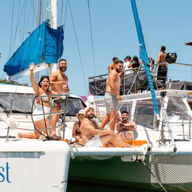 A group of smiling adults, some waving, enjoy a sunny day on sailboats. They are lounging on the decks with clear skies and water in the background. The boat's name "Catalyst" is visible, along with logos for "Sail Gustavo" and "Capt. Josh's Excursions.