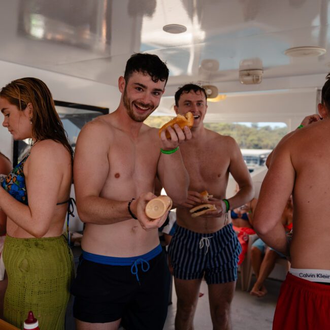 A group of people in swimsuits are gathered on a boat, with one person in the center holding food and smiling at the camera. Others appear to be preparing or eating food, enjoying a luxury yacht hire Sydney experience on a beautiful day.