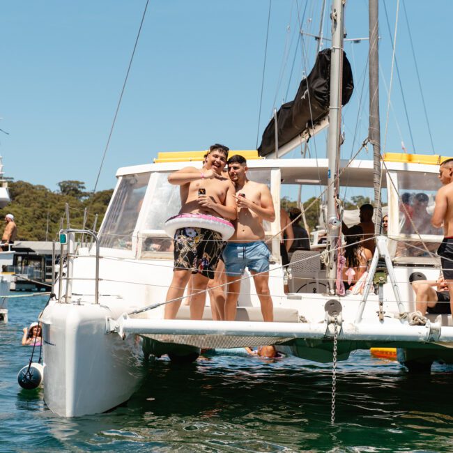 A group of people enjoying a sunny day on a sailboat. Some are standing and posing for a picture, while others are lounging and relaxing with drinks. Another nearby boat in the background. Water and greenery visible around them. The atmosphere is lively and festive.