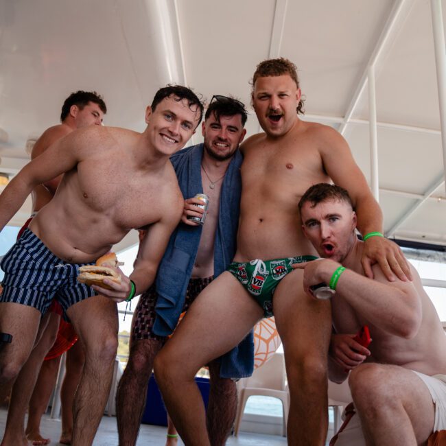 A group of five men in swimwear pose together on a boat. One holds a snack, and another holds a beverage. They appear to be enjoying themselves during what seems like a luxury yacht hire Sydney experience.