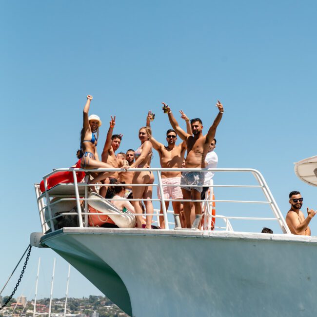 A group of people in vibrant swimwear stands on the deck of a yacht, smiling and raising their hands in celebration. The yacht glides on a calm body of water under a clear, sunny sky, with a picturesque shoreline and buildings visible in the background. Logos subtly appear in the bottom corners.
