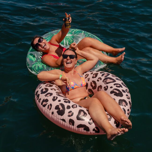 Two women in swimsuits float on inflatable rings in a body of water during a private yacht charter on Sydney Harbour. One woman holds a drink can, and both smile and pose for the camera.