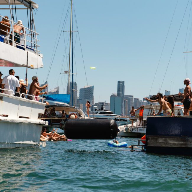 People are enjoying a sunny day on the water with multiple boats docked close together. Some are on the decks while others swim or float in the water. The skyline of a city with tall buildings is visible in the background, creating a picturesque scene. Logos for "Sail Gustavo" and "Latino Seeds" add to the charm.