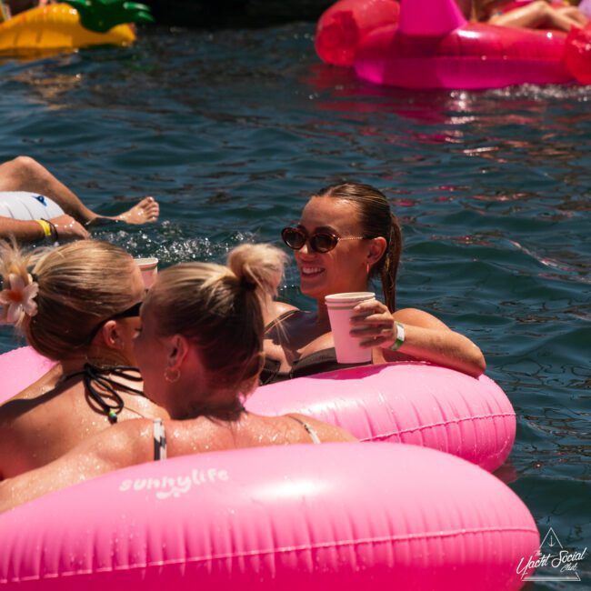 People relax on pink inflatable rafts in a body of water, holding drinks. Others in the background enjoy various inflatables, making it feel like a catamaran party in Sydney Harbour.