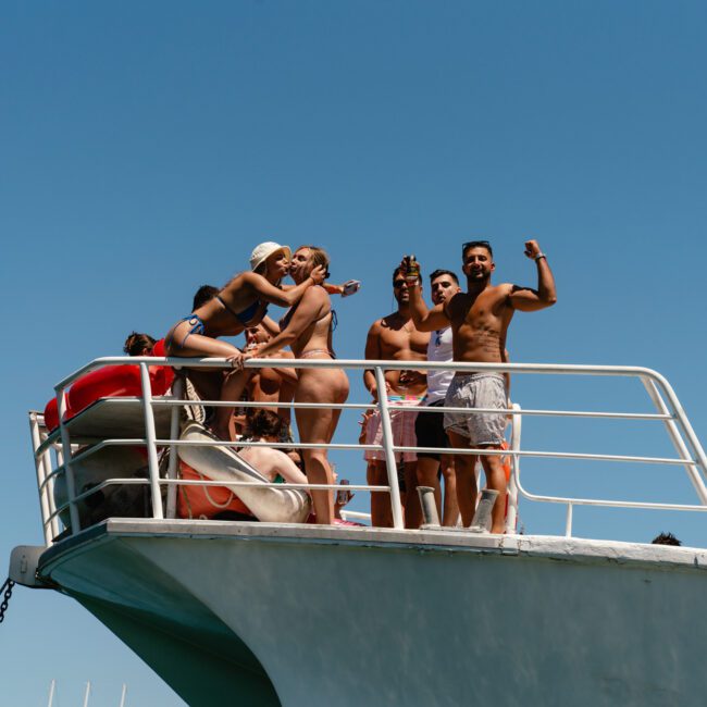 A group of young adults in swimwear are gathered on the deck of a boat, some posing with their arms raised and flexing. The sky is clear and blue. The boat has logos that read "Sail Gustavo" and "The Yacht Social Club," adding a touch of sophistication to their sun-soaked fun.