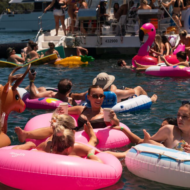 People are enjoying a sunny day on the water with inflatable pool floats near a boat. Various floats include a pink flamingo and a brown deer. Some individuals are holding drinks, adding to the festive atmosphere of their Sydney boat party hire.