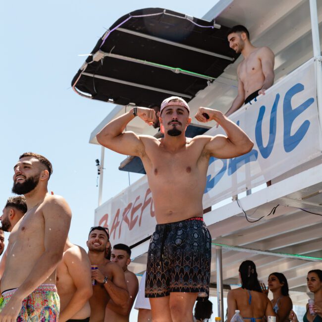 A man in swim trunks flexes his muscles while standing on a platform at a beach party. Other people in swimwear are around him, enjoying the lively and festive atmosphere. A sign behind him reads "Barefoot Blue," and the "Sal Gustavo" logo is visible, encapsulating the essence of summer fun.