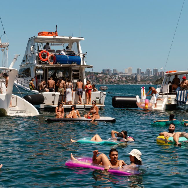A lively scene of people enjoying a sunny day on the water, several groups floating on inflatables, swimming, or lounging near two anchored boats. The city skyline is visible in the background under a clear blue sky, with the vibrant atmosphere creating a sense of cheerful relaxation.