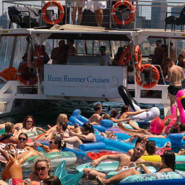 A group of people enjoy a sunny day on inflatable floats in the water next to a boat with a "Rum Runner Cruises" banner. The boat, crowded with happy guests, features the Yacht Social banner and offers an exclusive private yacht charter on Sydney Harbour.