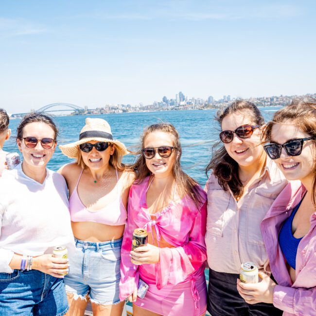 A group of people stands on a boat with drinks in hand, smiling at the camera. The background features a body of water and a city skyline with the Sydney Harbour Bridge in view, perfect for a memorable catamaran party in Sydney.