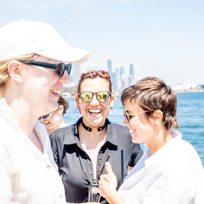 Three people smile and laugh together outdoors near a body of water, with part of a city skyline and a bridge visible in the background, perhaps enjoying a Sydney boat party hire.