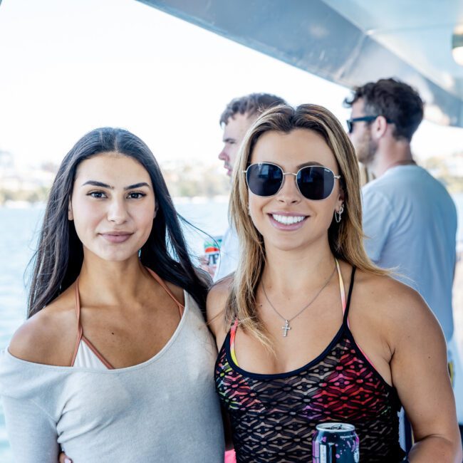 Two women smiling on a boat with water and cityscape in the background, surrounded by other people. One wears sunglasses and a cross necklace, the other wears a light-colored top. They seem to be enjoying a catamaran party in Sydney.