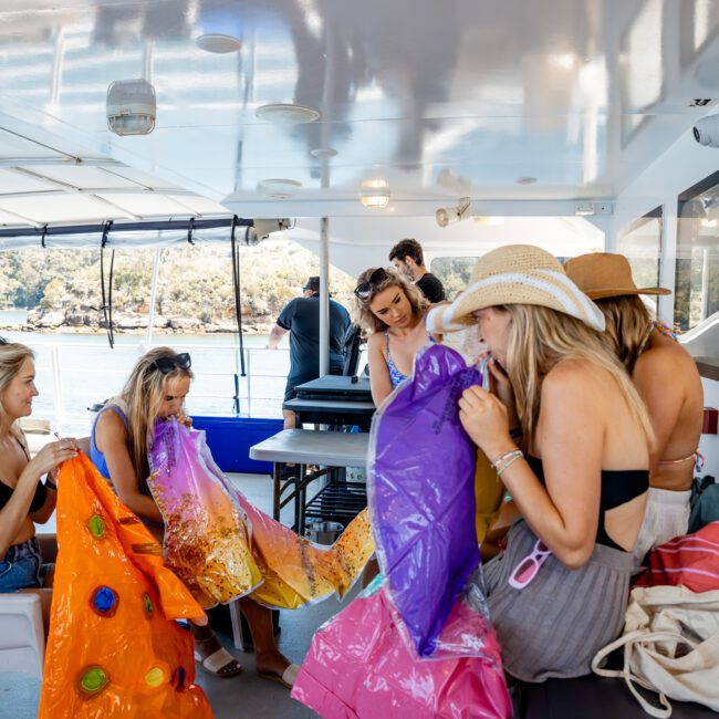 A group of people on a boat are inflating colorful pool floats. The luxury yacht hire in Sydney features open sides and white walls, with everyone dressed in swimwear.