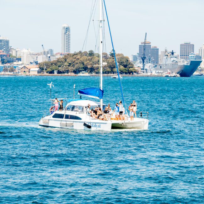 A catamaran with several people on board sails in calm waters with a city skyline and a naval ship in the background, perfect for a Sydney boat party hire.