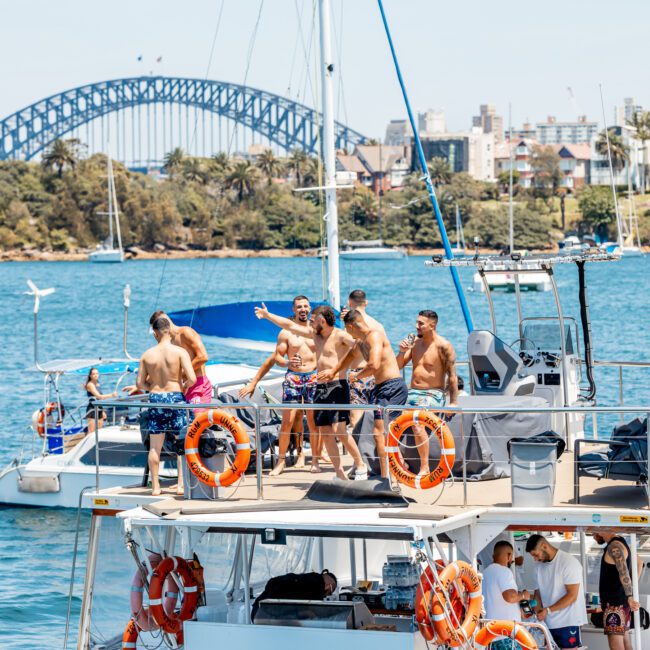 A group of men on a boat enjoy a sunny day on the water, with the Sydney Harbour Bridge visible in the background — a perfect setting for a Sydney boat party hire.