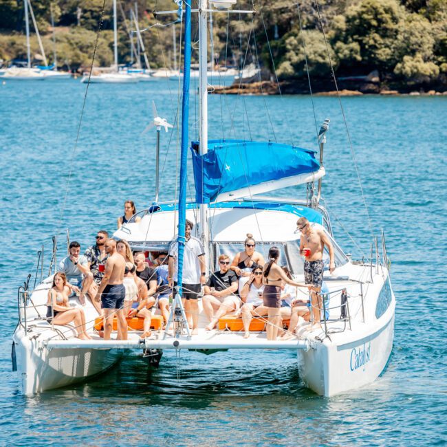 A group of people relax and socialize on a sailboat in a calm body of water, with several other boats and a green shoreline in the background, enjoying their private yacht charter on Sydney Harbour.