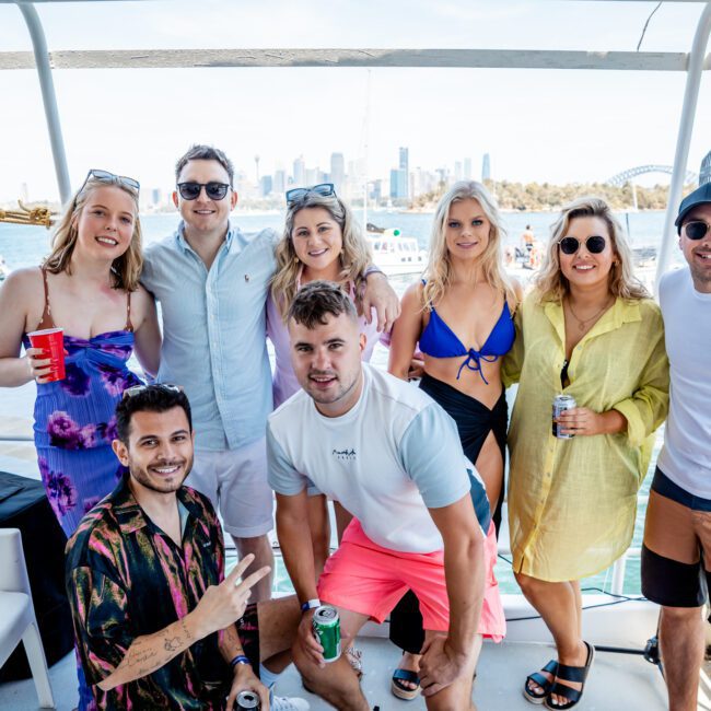 A group of eight people smiling and posing together on a boat with a city skyline and water in the background. They are dressed in casual summer attire, some holding drinks. It's clear they're enjoying a Catamaran party Sydney has to offer, perfect for any Corporate boat events Sydney hosts throughout the season.