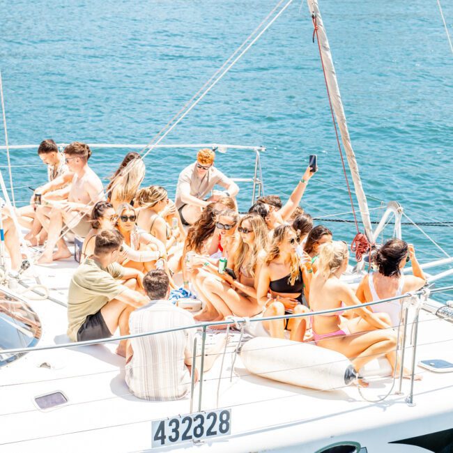 A group of people is sitting and standing on the deck of a sailboat on clear blue water near a rocky shoreline, enjoying a lively catamaran party in Sydney Harbour.