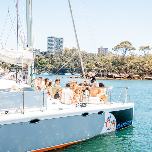 A group of people are gathered on the deck of a white sailboat with a logo, sailing in a calm body of water near a wooded shoreline and several tall buildings in the background, enjoying their private yacht charter Sydney Harbour.