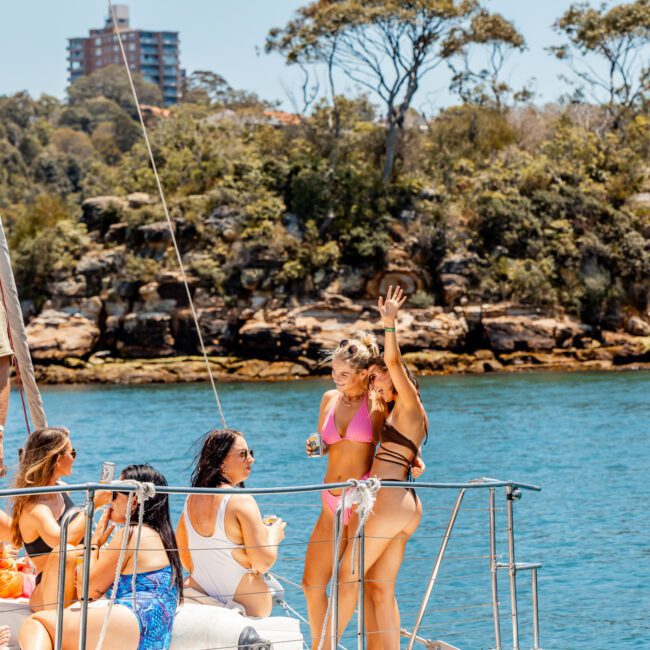 A group of people enjoy a sunny day on a luxury yacht hire in Sydney, with lush trees and cliffs in the background. Some are drinking beverages, and one woman in a pink bikini is waving.