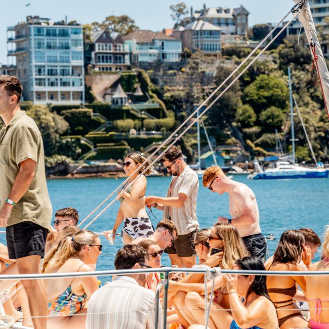 A group of people in swimwear socializing on a boat with coastal homes and trees visible in the background on a sunny day, perfect for a Sydney boat party hire.