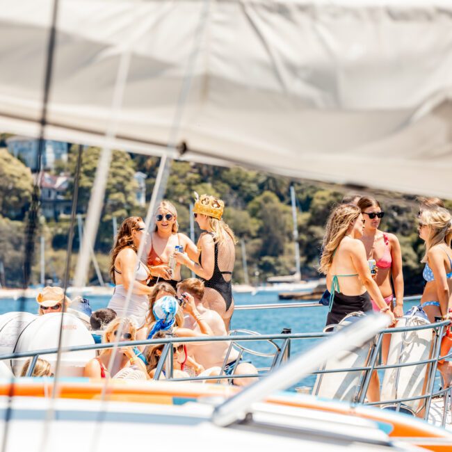 A group of people socialize on a luxury yacht hire in Sydney under a canopy on a sunny day, with a scenic background featuring trees and water. Some are sitting, others are standing, and many are wearing swimwear.