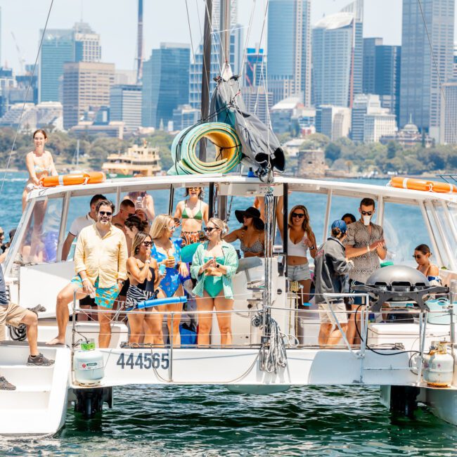 A group of people in swimwear are gathered on a private yacht charter in Sydney Harbour with a city skyline in the background. The boat is equipped with a grill and water hoses on the deck, perfect for a fun Sydney boat party hire.