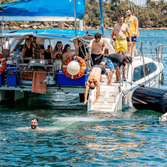 People are enjoying a sunny day on a catamaran in clear blue waters. Some are diving or swimming in the water, while others relax onboard under a blue canopy during their Sydney boat party hire.