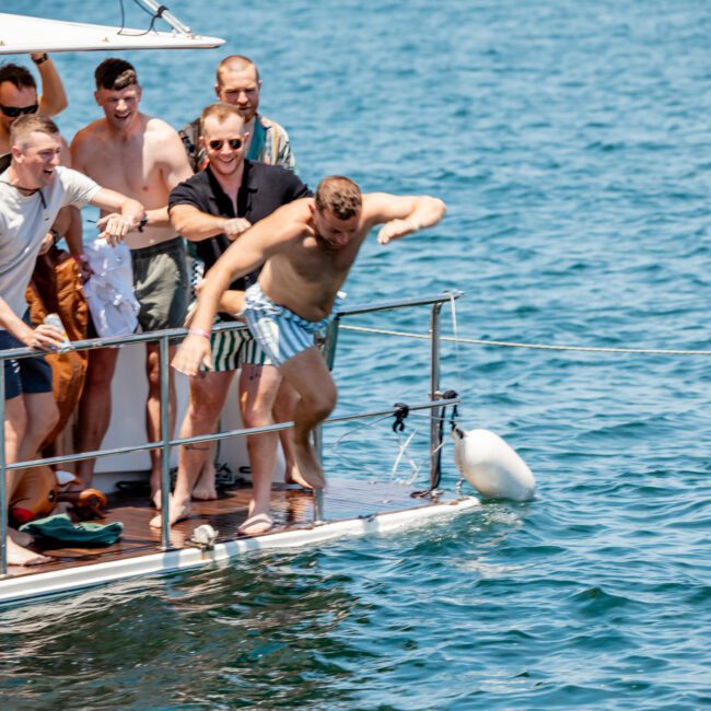 A group of men on a boat watch one of them jump into the water, while others smile and cheer. They are all casually dressed, enjoying a sunny day on a lake or the ocean. This could easily be scenes from a Sydney boat party hire or a private yacht charter on Sydney Harbour.