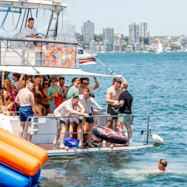 A group of people are gathered on a luxury yacht hire in Sydney, some in swimwear, with an inflatable raft floating nearby in the water. The yacht is on a sunny body of water, with a stunning city skyline in the background.