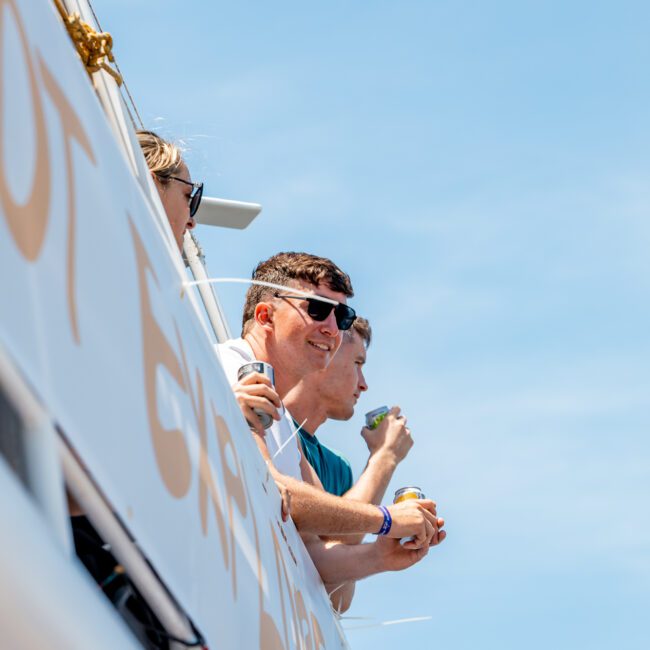 Three people wearing sunglasses are standing on the deck of a catamaran, holding drinks, and looking out towards the water under a clear blue sky.