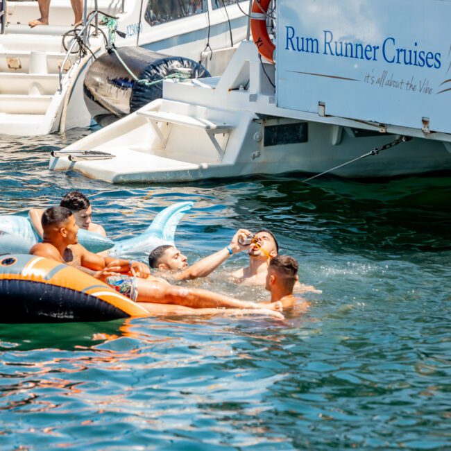 Four people are relaxing on an inflatable in the water next to a boat labeled "Rum Runner Cruises." They are enjoying drinks and the sunny weather during a Sydney boat party hire.