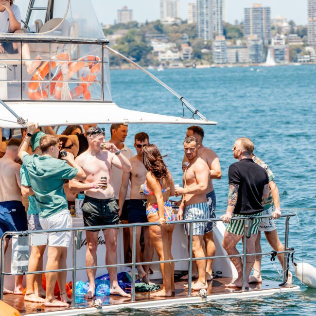 A lively group enjoying drinks on a sunny day aboard a private yacht charter near the Sydney Harbour shoreline with buildings in the background.