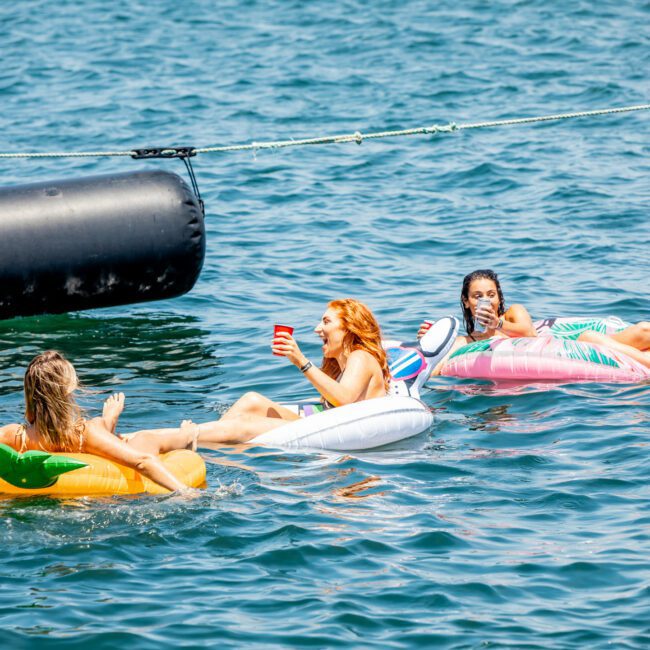 Three individuals float on colorful inflatables in the water, enjoying a sunny day. One person holds a drink while others relax nearby. A large luxury yacht hire Sydney is seen in the background, adding an elegant touch to the scene.