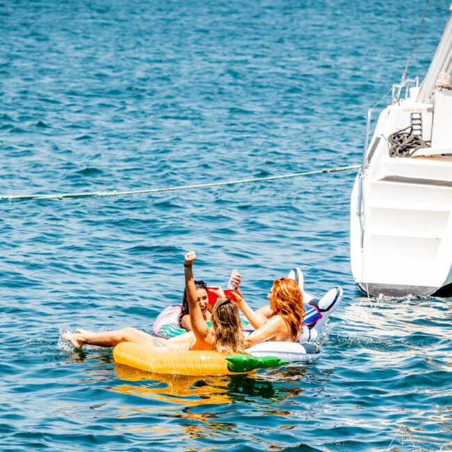 Three people are floating on inflatable toys, including a unicorn and pineapple, in the water near a luxury yacht hire Sydney on a sunny day.